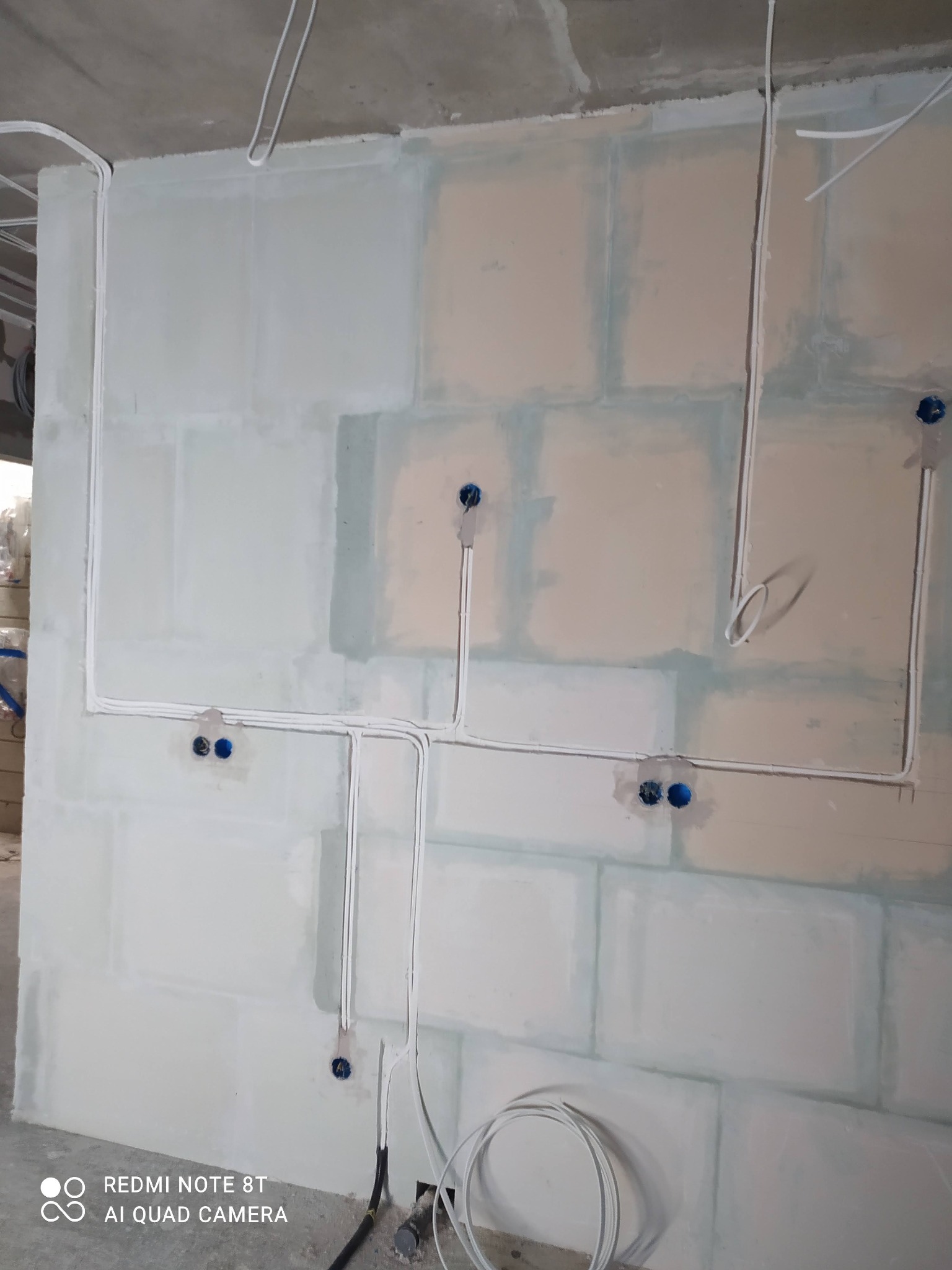 Świeżo położone przewody elektryczne w białych rurkach na ścianie z płyt kartonowo-gipsowych, widoczne niebieskie puszki pod gniazdka, surowy stan wykończenia wnętrza.