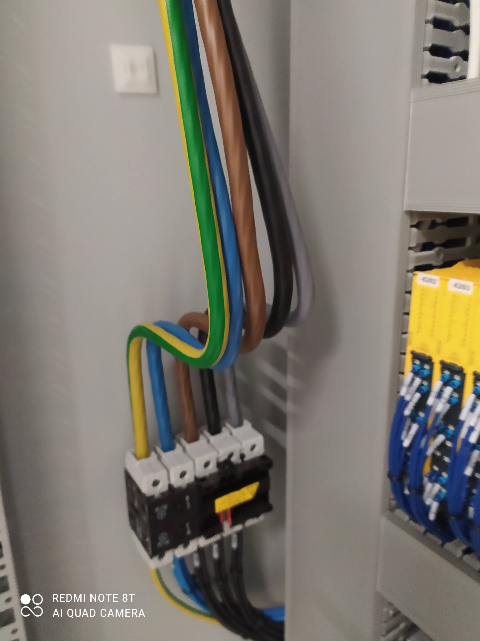 Zbliżenie na rozdzielnicę elektryczną z kolorowymi przewodami (żółto-zielony, niebieski, brązowy, szary) podłączonymi do wyłączników, widoczne także elementy montażowe i kable w listwach po prawej...