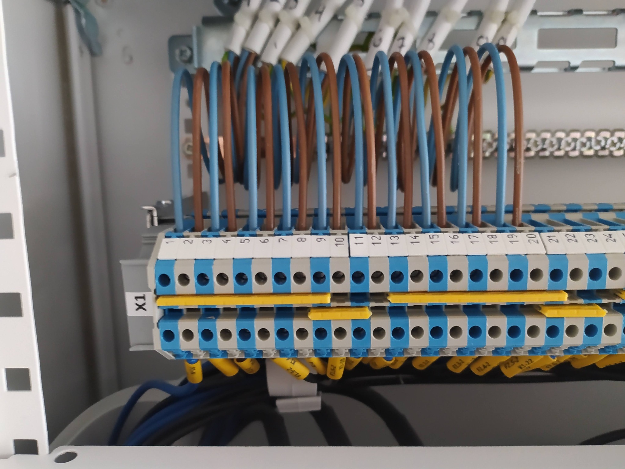 Szczegółowe ujęcie rozdzielnicy elektrycznej z podłączonymi przewodami w kolorach brązowym i niebieskim, oznaczonymi numerami na listwie zaciskowej, widoczne żółte zworki i oznaczenia przewodów.