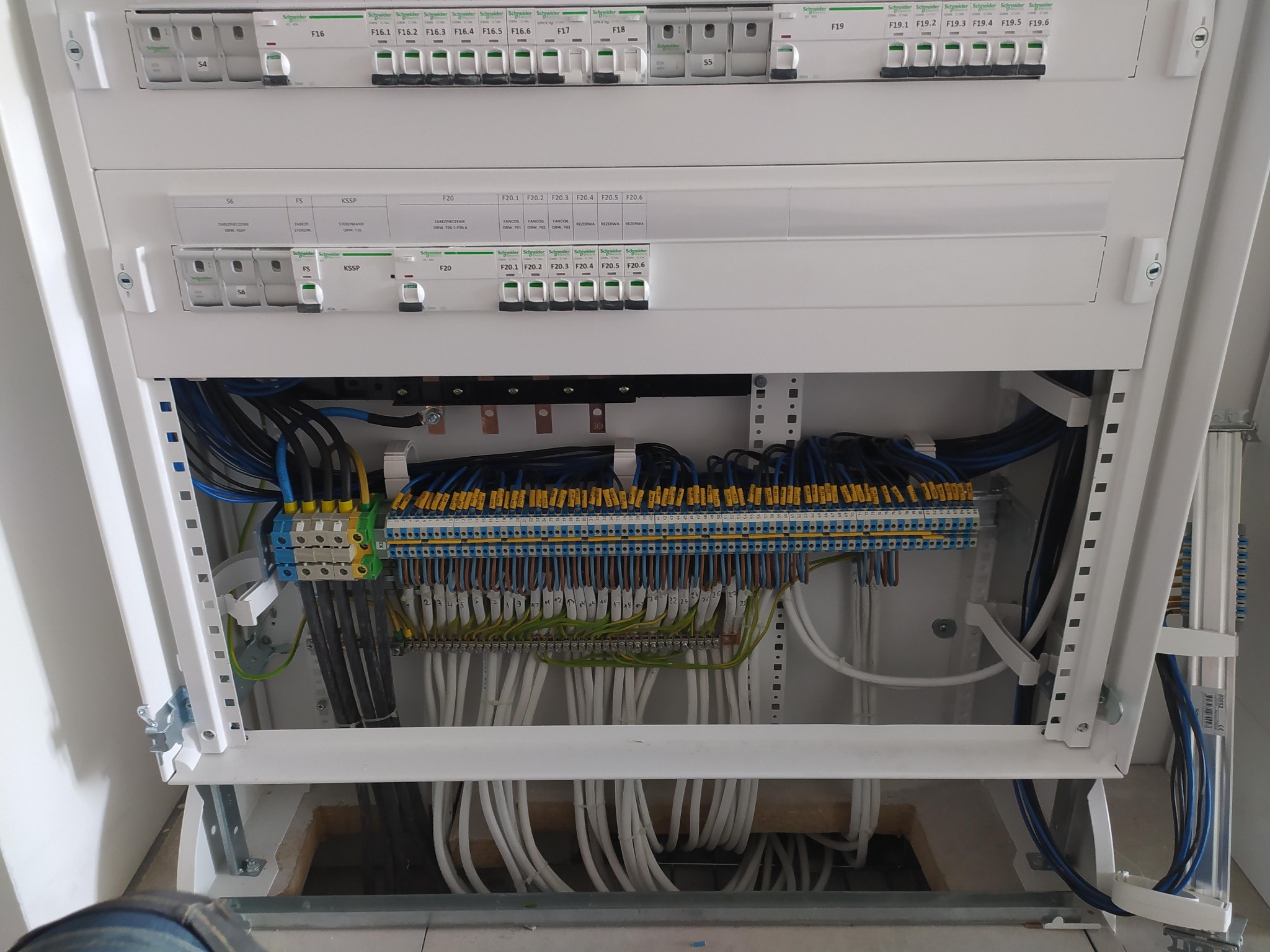 Otwarta, biała skrzynka rozdzielcza z licznymi bezpiecznikami Schneider Electric i starannie ułożonymi przewodami w różnych kolorach, zamontowana w pomieszczeniu z widocznym fragmentem podłogi...