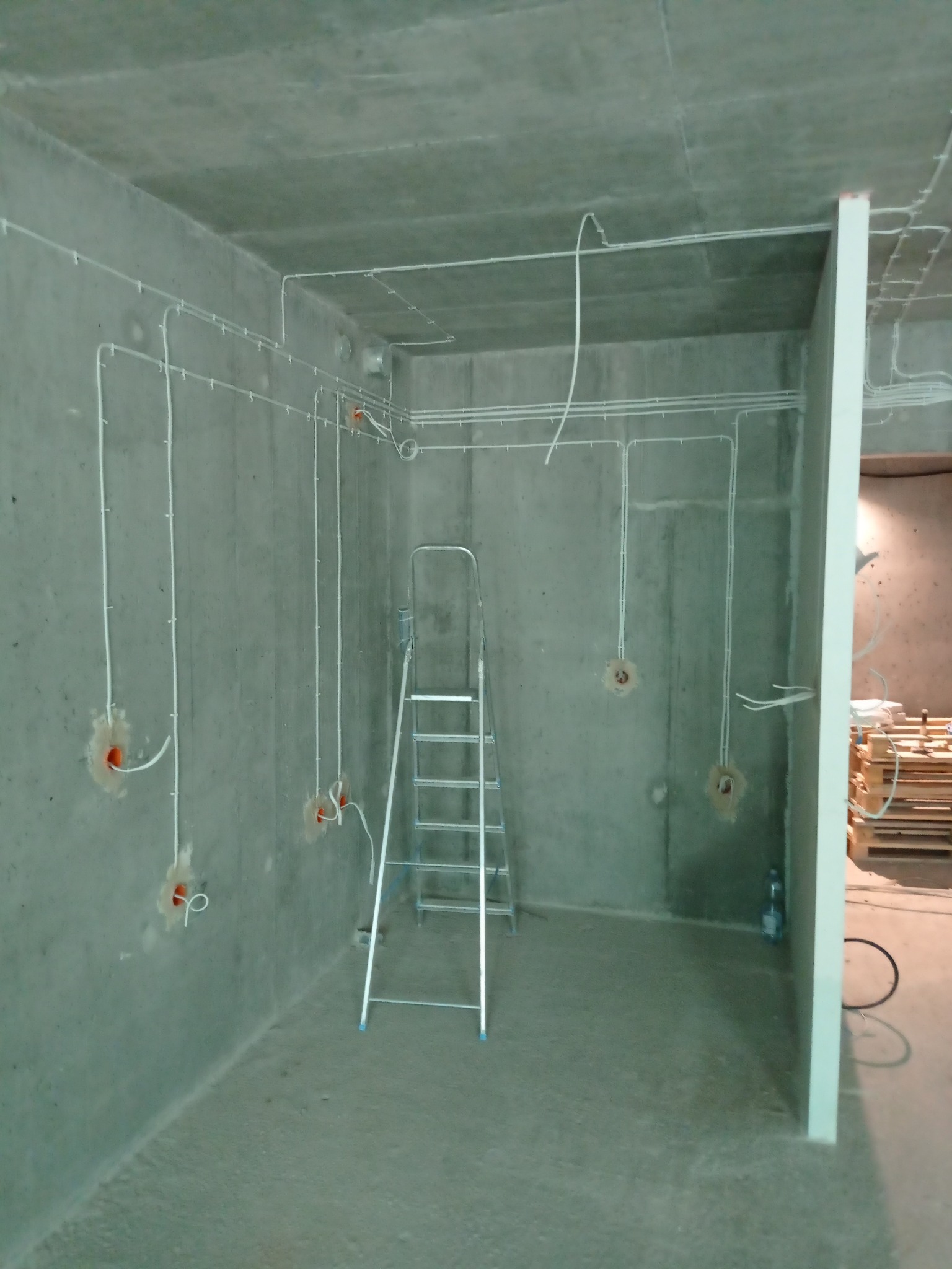 Surowy betonowy pokój w trakcie prac elektrycznych: białe przewody rozprowadzone po ścianach i suficie, otwory pod gniazdka, aluminiowa drabina oparta o ścianę, butelka wody przy ścianie.