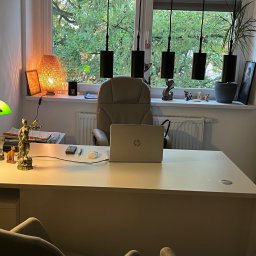 KANCELARIA ADWOKACKA ADWOKAT DOROTA WYPUSZ-KUCH - Biuro z białym biurkiem, laptopem, lampką z zielonym kloszem i figurką Temidy, widok na zielone drzewa za oknem.