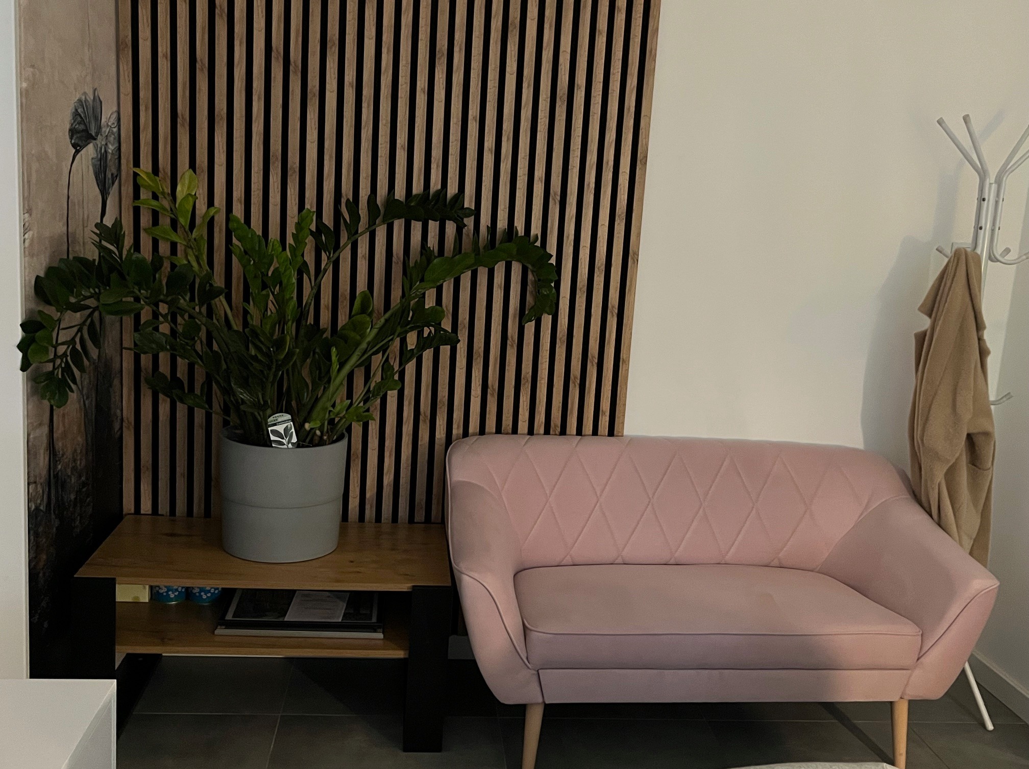 Jasnoróżowa sofa z pikowaniem w romby, drewniany stolik z rośliną doniczkową, dekoracyjna ściana z drewnianych listewek i wieszak na ubrania w tle.