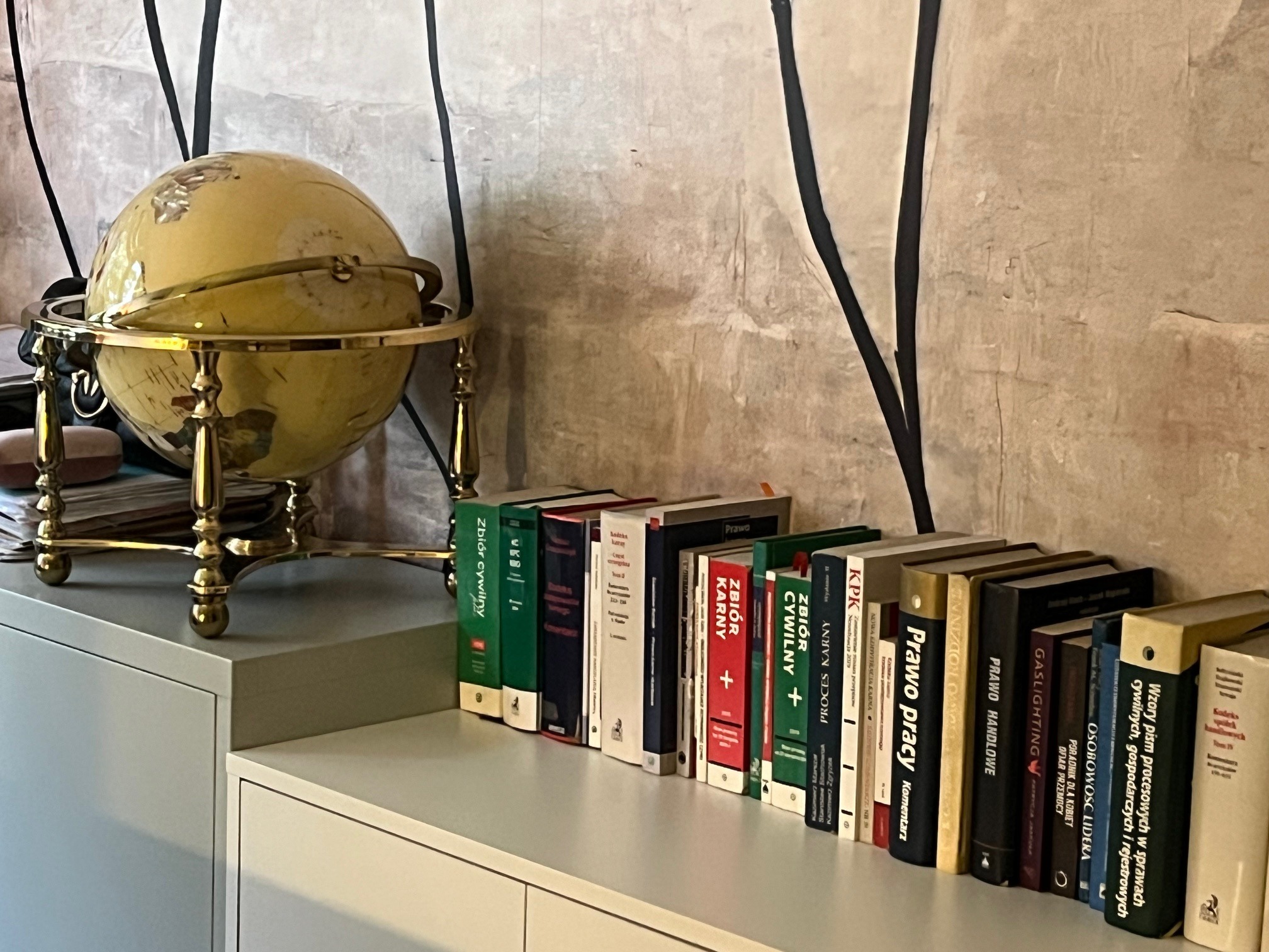 Globus na szafce obok rzędu książek prawniczych, w tym 'Prawo Pracy', 'Zbiór Cywilny', 'Zbiór Karny', 'Prawo Handlowe' i 'KPK', na tle ściany z abstrakcyjnym wzorem.