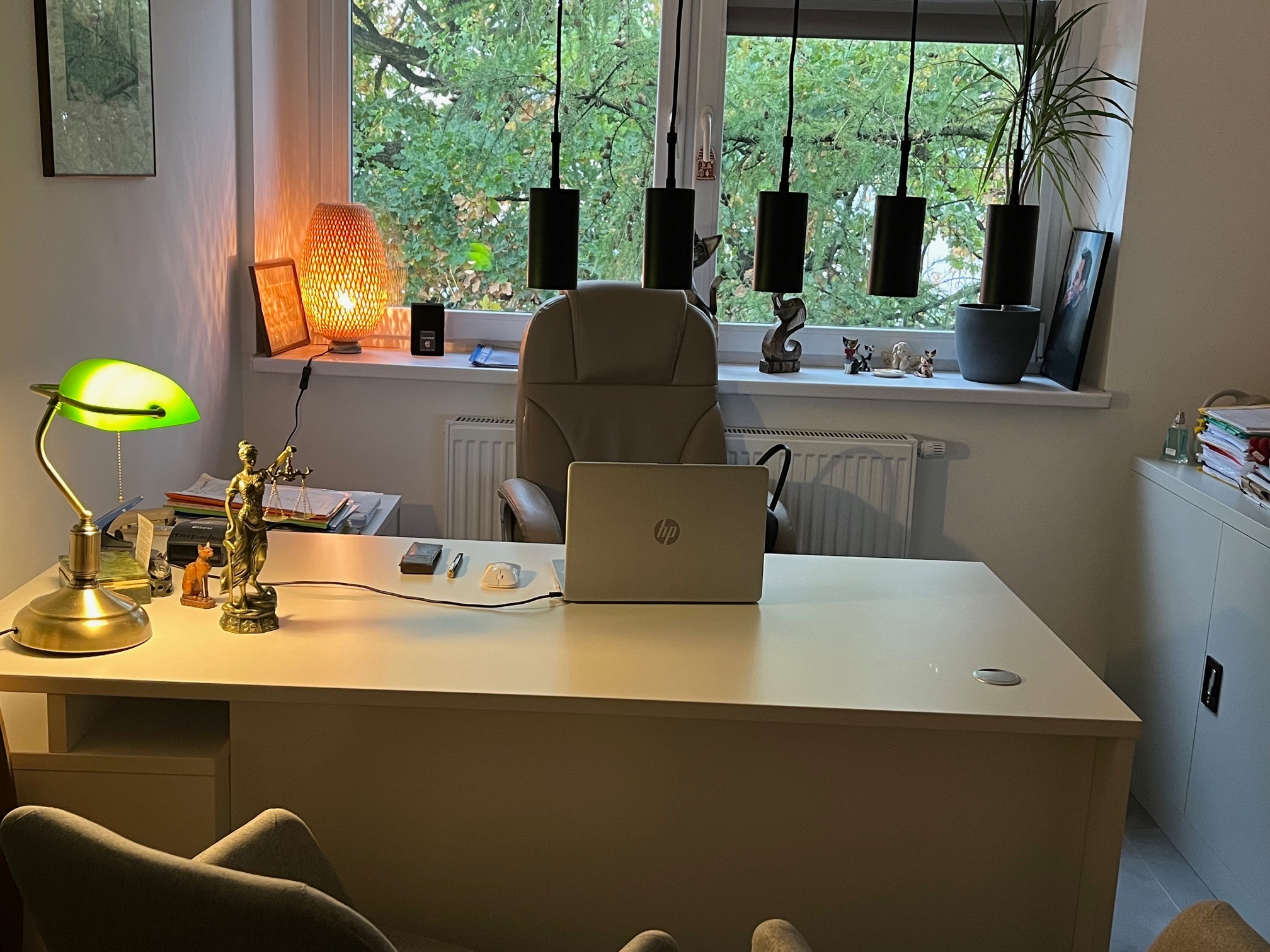Biuro z białym biurkiem, laptopem, lampką z zielonym kloszem i figurką Temidy, widok na zielone drzewa za oknem.