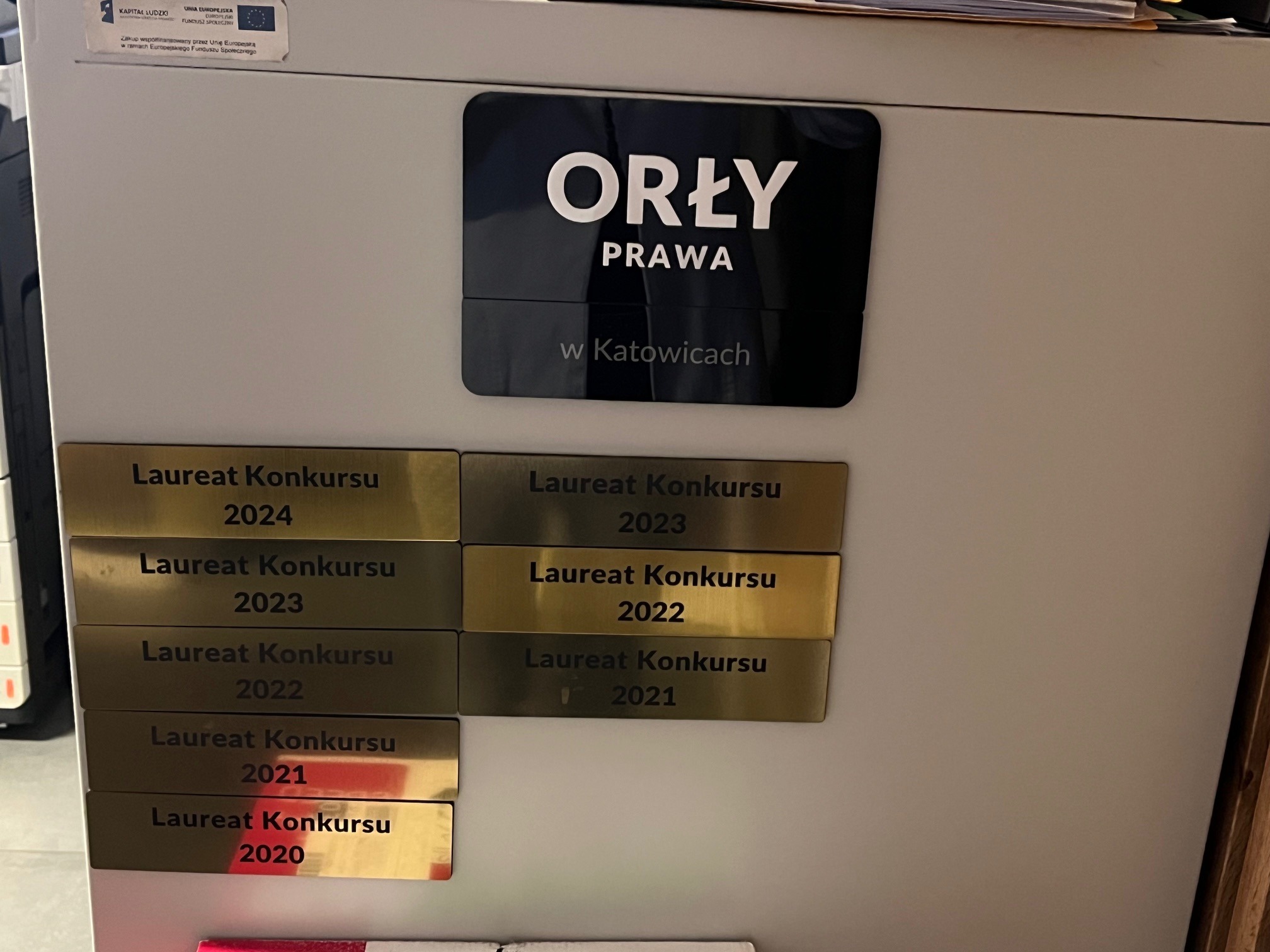 Tablica z logo 'Orły Prawa w Katowicach' i złotymi tabliczkami 'Laureat Konkursu' z lat 2020-2024, umieszczona na szafce biurowej.