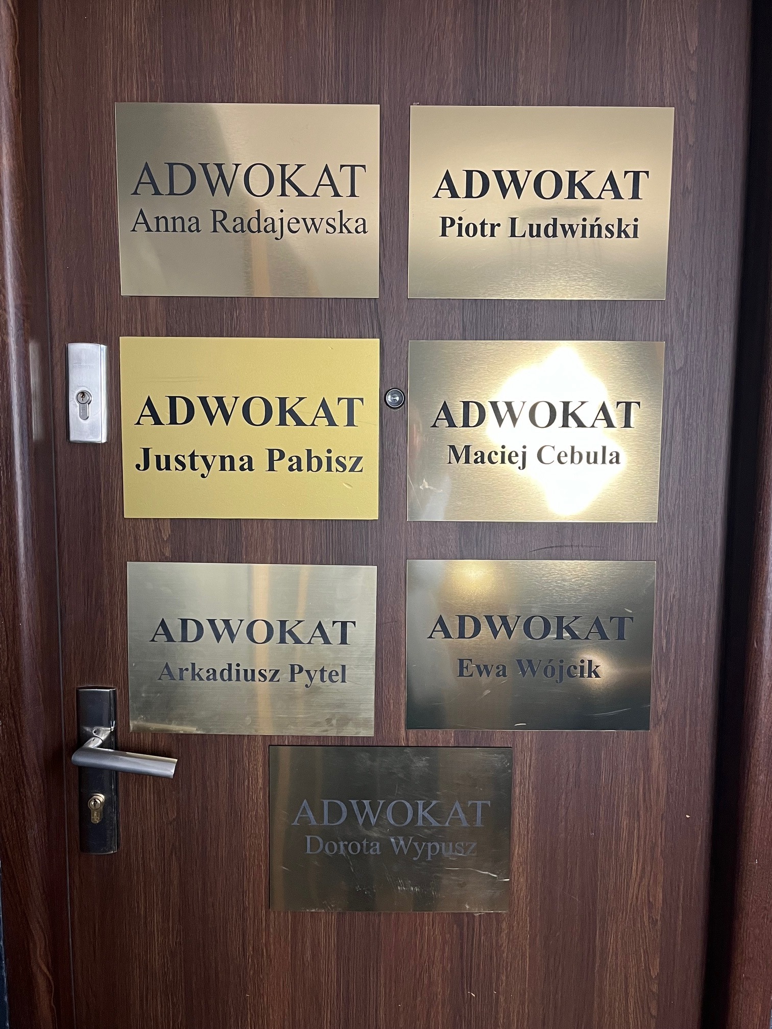 Tabliczki z nazwiskami i tytułem 'Adwokat' umieszczone na drzwiach, w różnych odcieniach złota i brązu.
