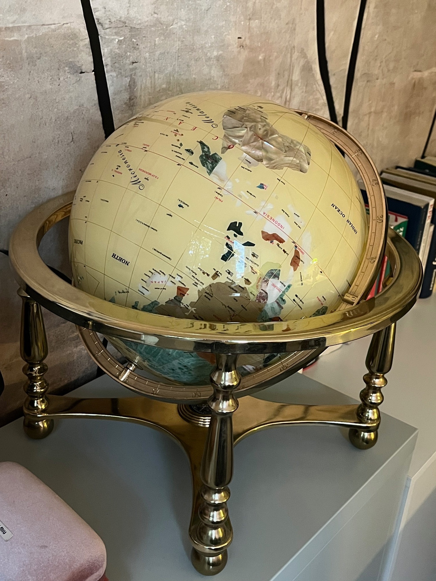 Antyczny globus z mosiężną podstawą i kremową mapą świata, stojący na szarym blacie, z książkami w tle i fragmentem ściany z ciemnymi pionowymi wzorami.