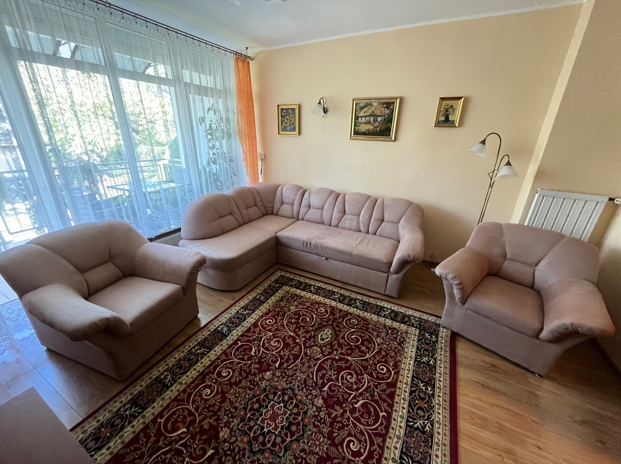 Jasny salon z narożną kanapą, dwoma fotelami i wzorzystym dywanem w odcieniach czerwieni i beżu. Na ścianie wiszą obrazy w złotych ramach, a obok okna z firankami stoi lampa.