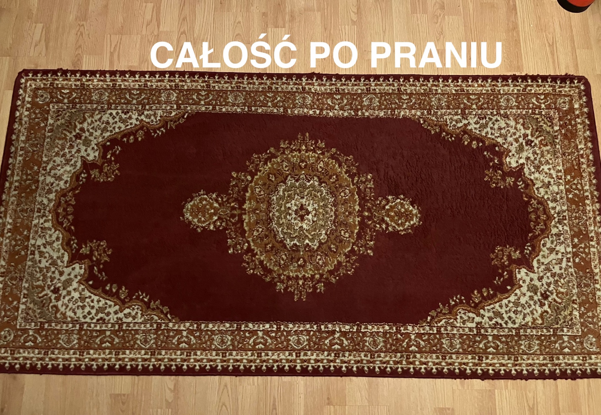 Czerwony dywan z orientalnym wzorem, leżący na drewnianej podłodze, sfotografowany z góry po procesie czyszczenia, z widocznym napisem 'CAŁOŚĆ PO PRANIU'.