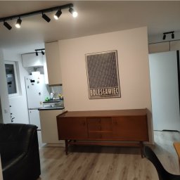 Midgard - Pokój z drewnianym kredensem pod plakatem Bolesławiec, widoczne oświetlenie punktowe na suficie i fragment kuchni z białą zabudową.