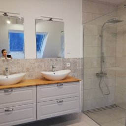Midgard - Nowoczesna łazienka z dwoma umywalkami nablatowymi, szarą szafką pod umywalki z drewnianym blatem, mozaiką nad szafką, kabiną prysznicową typu walk-in oraz osobą fotografującą wnętrze widoczną...