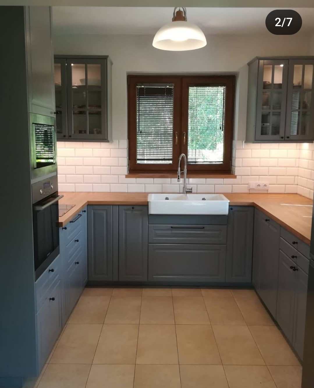 Szara kuchnia w stylu klasycznym z białym ceramicznym zlewem, drewnianym blatem i białymi płytkami na ścianie, widok na okno z żaluzjami.