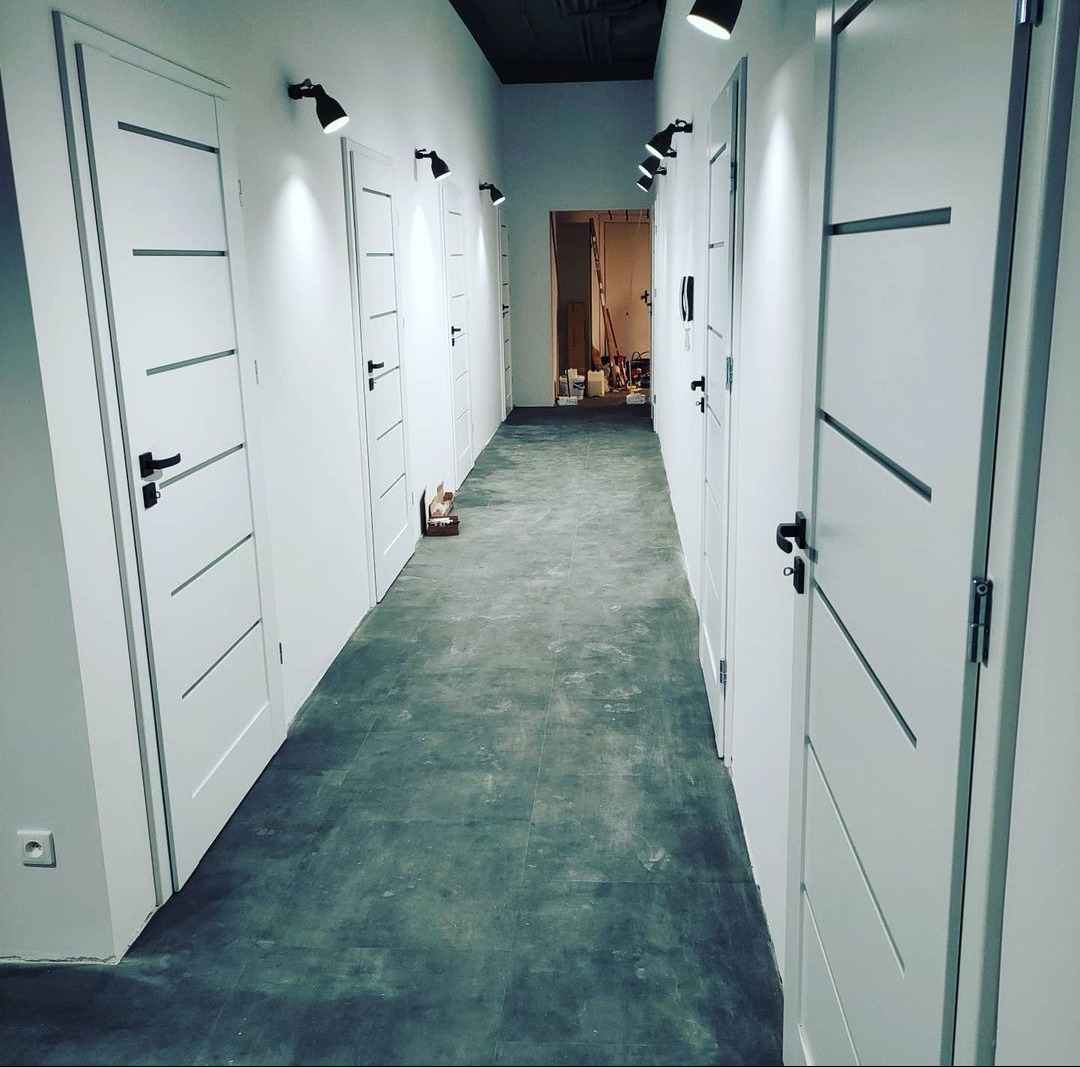 Długi korytarz z rzędem białych drzwi z czarnymi klamkami i poziomymi wstawkami, szara podłoga imitująca beton, czarne reflektory sufitowe, widoczne ślady prac wykończeniowych w tle.