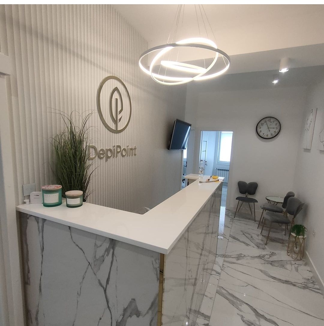 Recepcja w salonie kosmetycznym z marmurowym blatem, dekoracyjną ścianą z logo DepiPoint, nowoczesnym oświetleniem i marmurową podłogą.