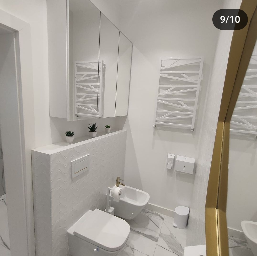 Biała łazienka z marmurową podłogą, toaletą, bidetem ze złotą armaturą, grzejnikiem drabinkowym i szafką z lustrami, udekorowana małymi sukulentami.