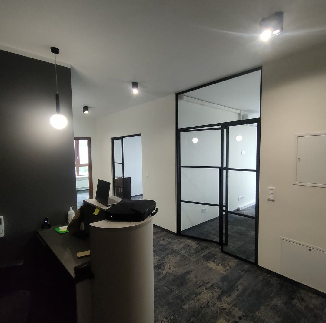 Nowoczesne biuro z czarnymi, przeszklonymi drzwiami działowymi i recepcją z laptopem, widok na korytarz z oknem.