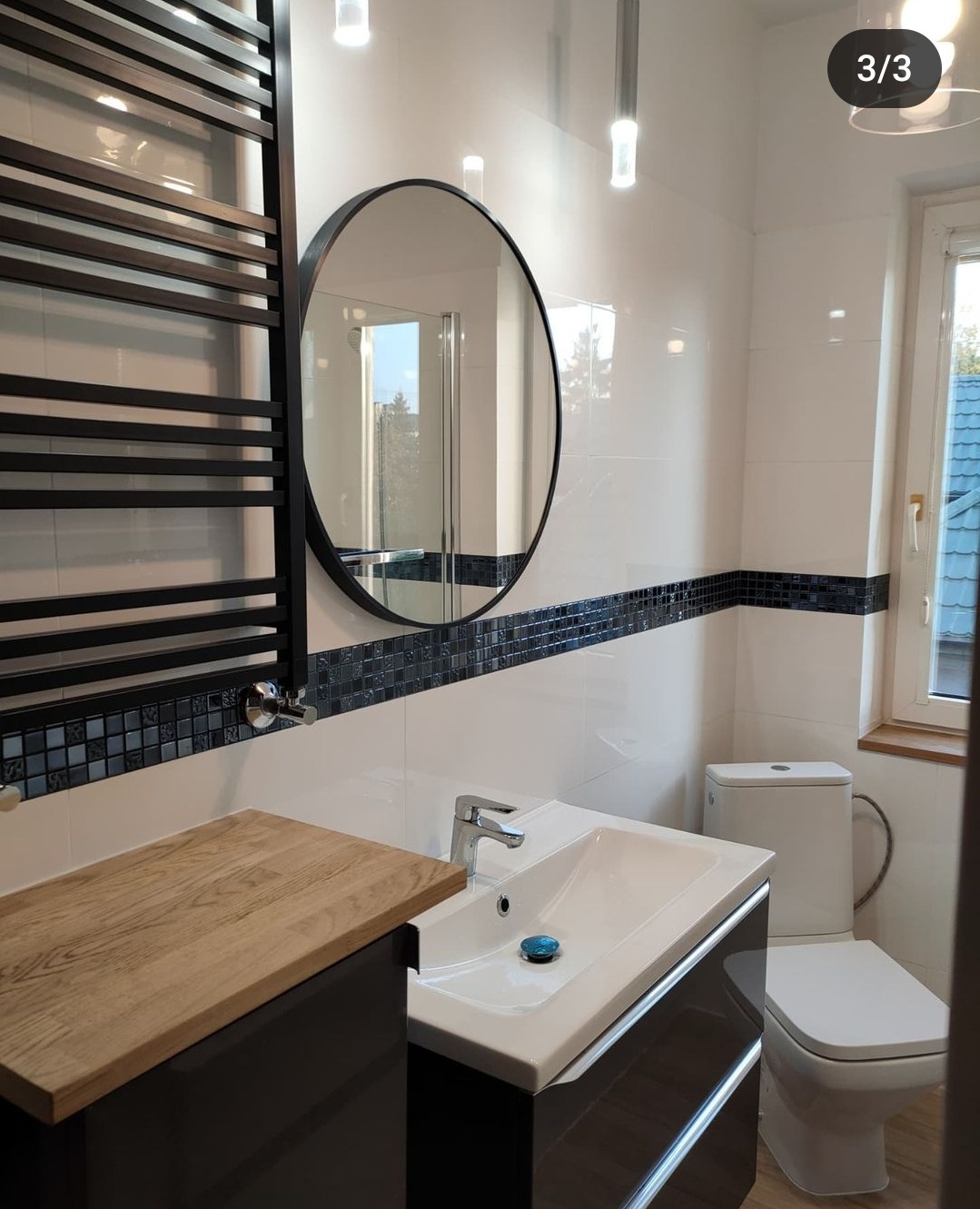 Nowoczesna łazienka z biało-czarnymi płytkami mozaikowymi, okrągłym lustrem w czarnej ramie, grzejnikiem drabinkowym i drewnianym blatem szafki umywalkowej.