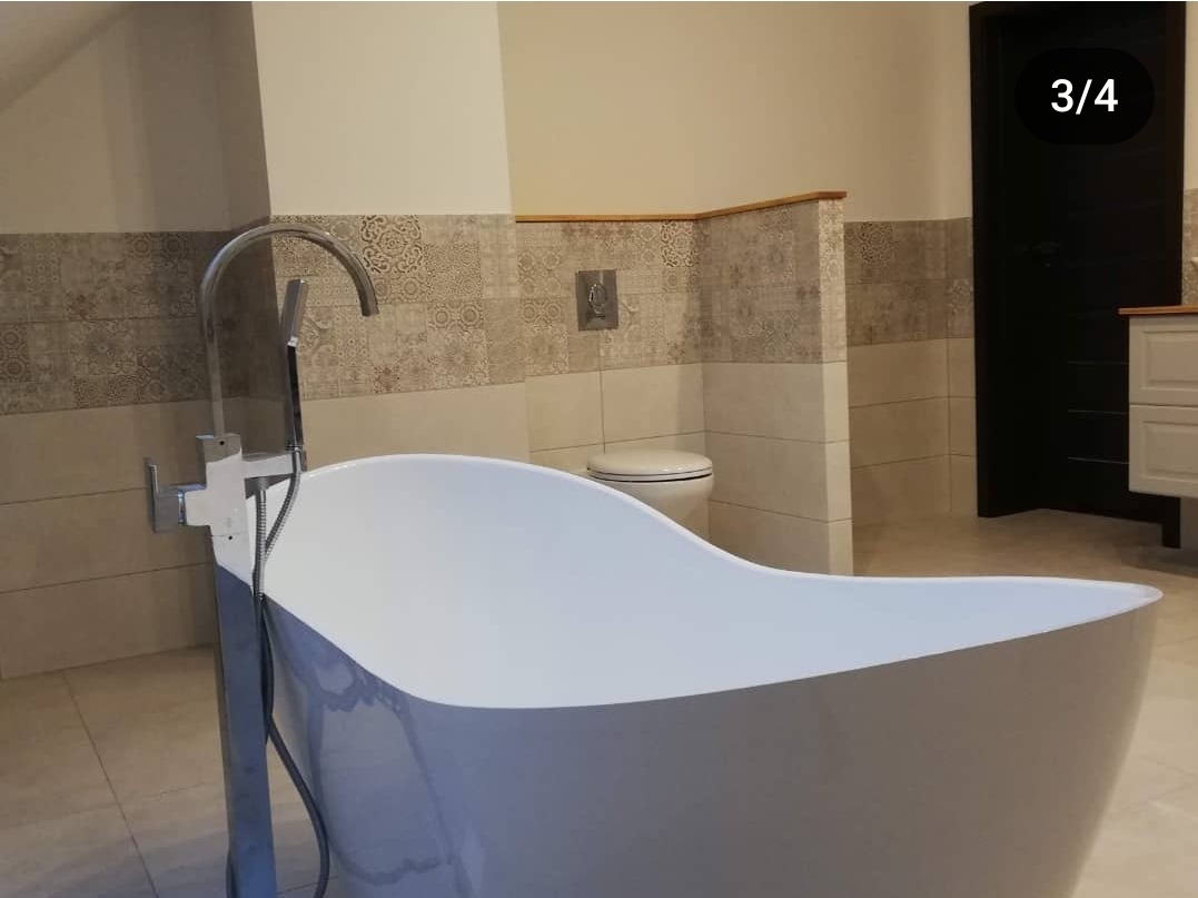 Wolnostojąca wanna z chromowaną baterią na tle wykończonej łazienki z mozaiką, widoczna toaleta i fragment szafki.