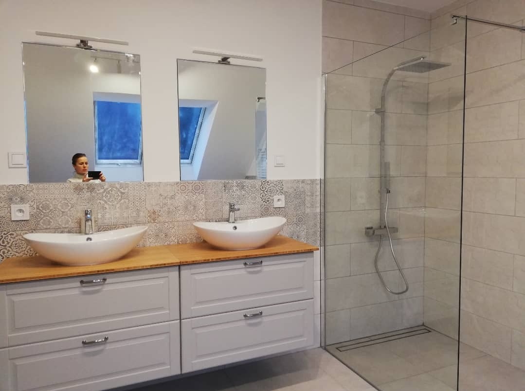 Nowoczesna łazienka z dwoma umywalkami nablatowymi, szarą szafką pod umywalki z drewnianym blatem, mozaiką nad szafką, kabiną prysznicową typu walk-in oraz osobą fotografującą wnętrze widoczną...