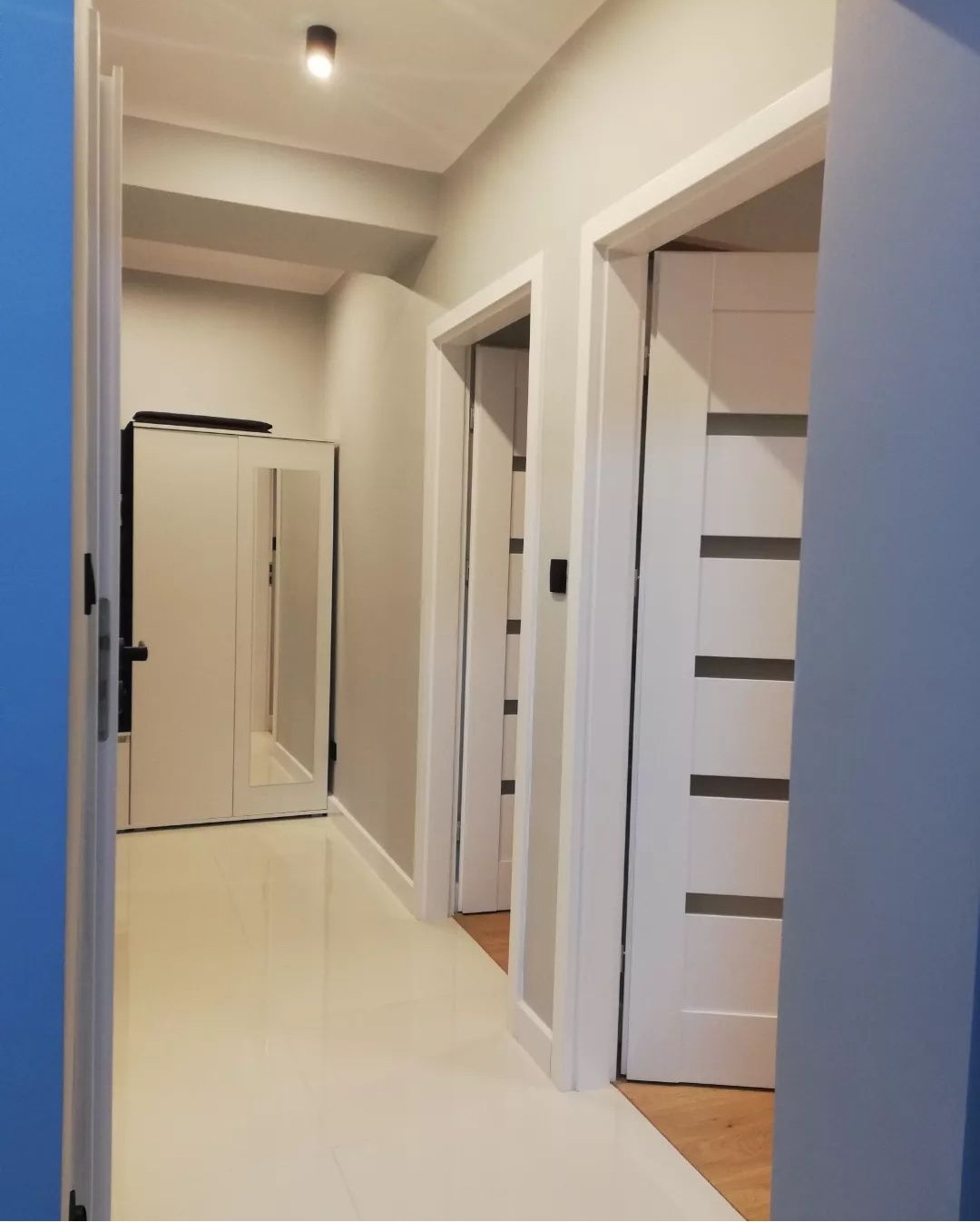 Korytarz po remoncie z białymi drzwiami z poziomymi przeszkleniami, jasnymi płytkami na podłodze i szafą z lustrem w tle.