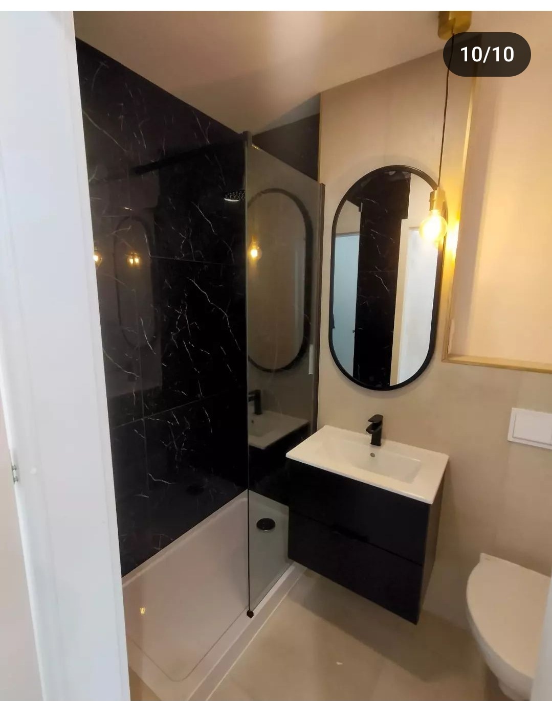 Nowoczesna łazienka z kabiną prysznicową typu walk-in, czarnymi płytkami imitującymi marmur, owalnym lustrem w czarnej ramie, wiszącą szafką z białą umywalką i złotą lampą wiszącą.
