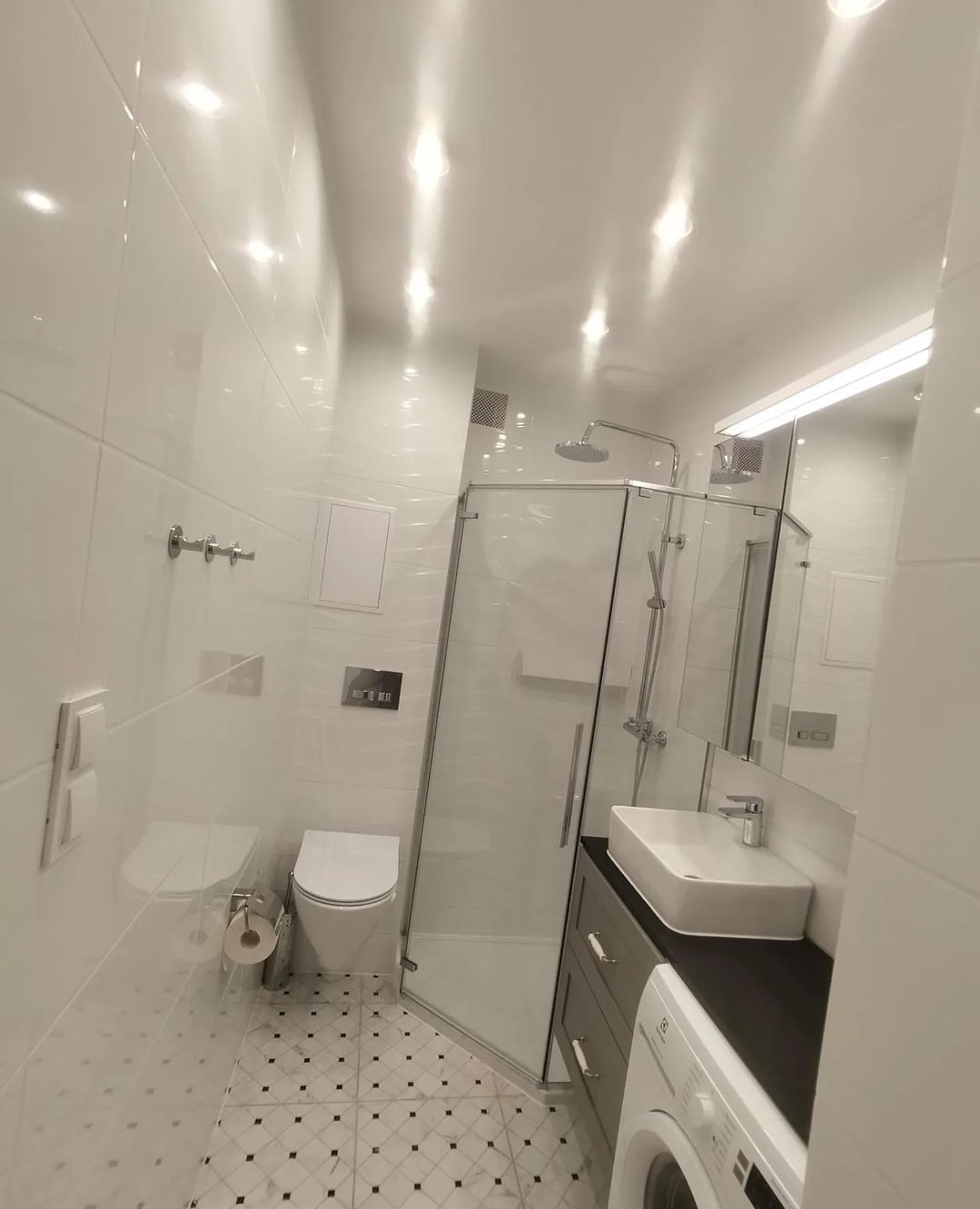 Nowoczesna łazienka z białą armaturą, kabiną prysznicową ze szklanymi ścianami, umywalką nablatową na ciemnej szafce i podłogą w biało-czarną szachownicę.