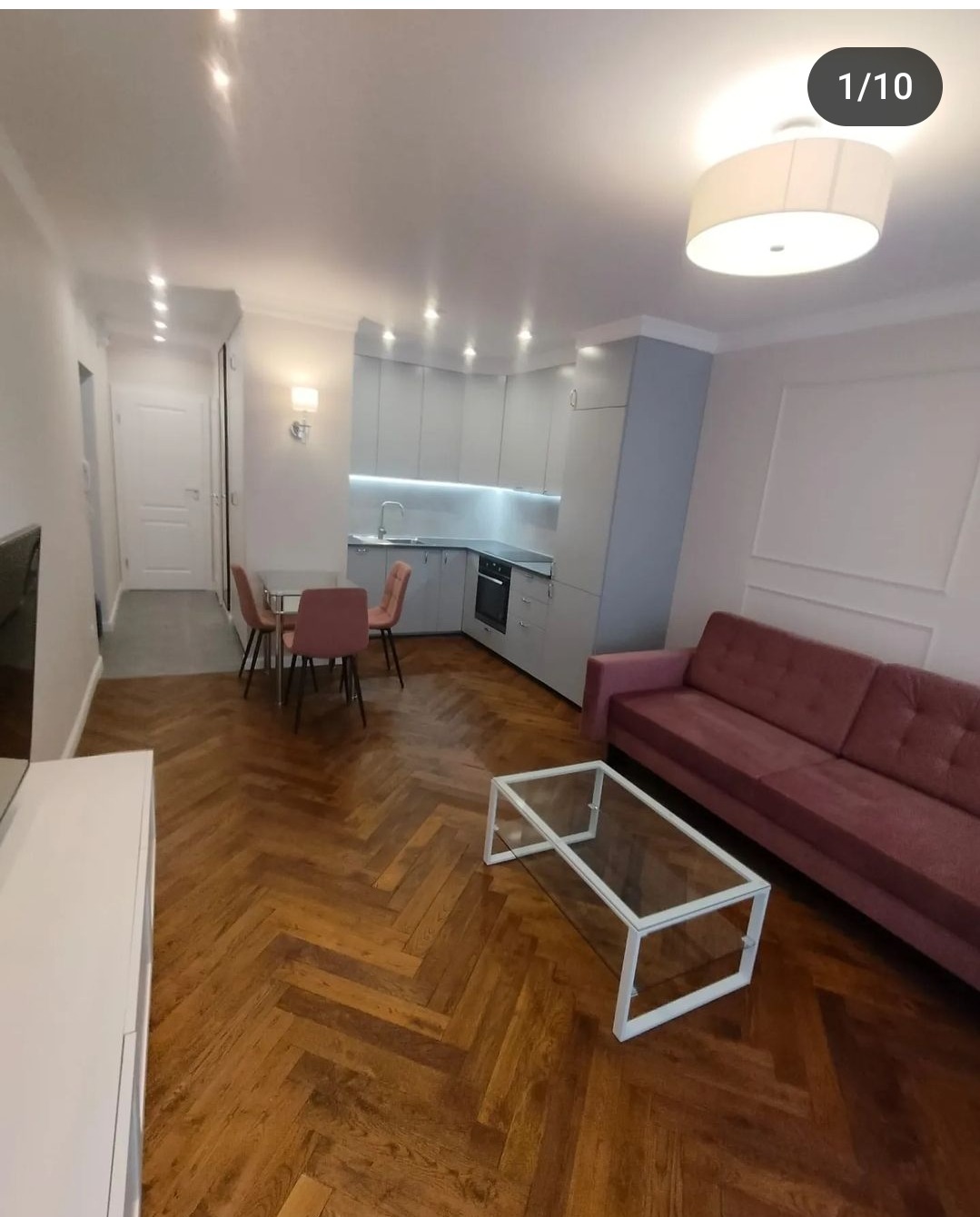 Jasne, nowocześnie urządzone wnętrze mieszkania z parkietem ułożonym w jodełkę, szarą kuchnią z podświetleniem LED, różową sofą i szklanym stolikiem kawowym.