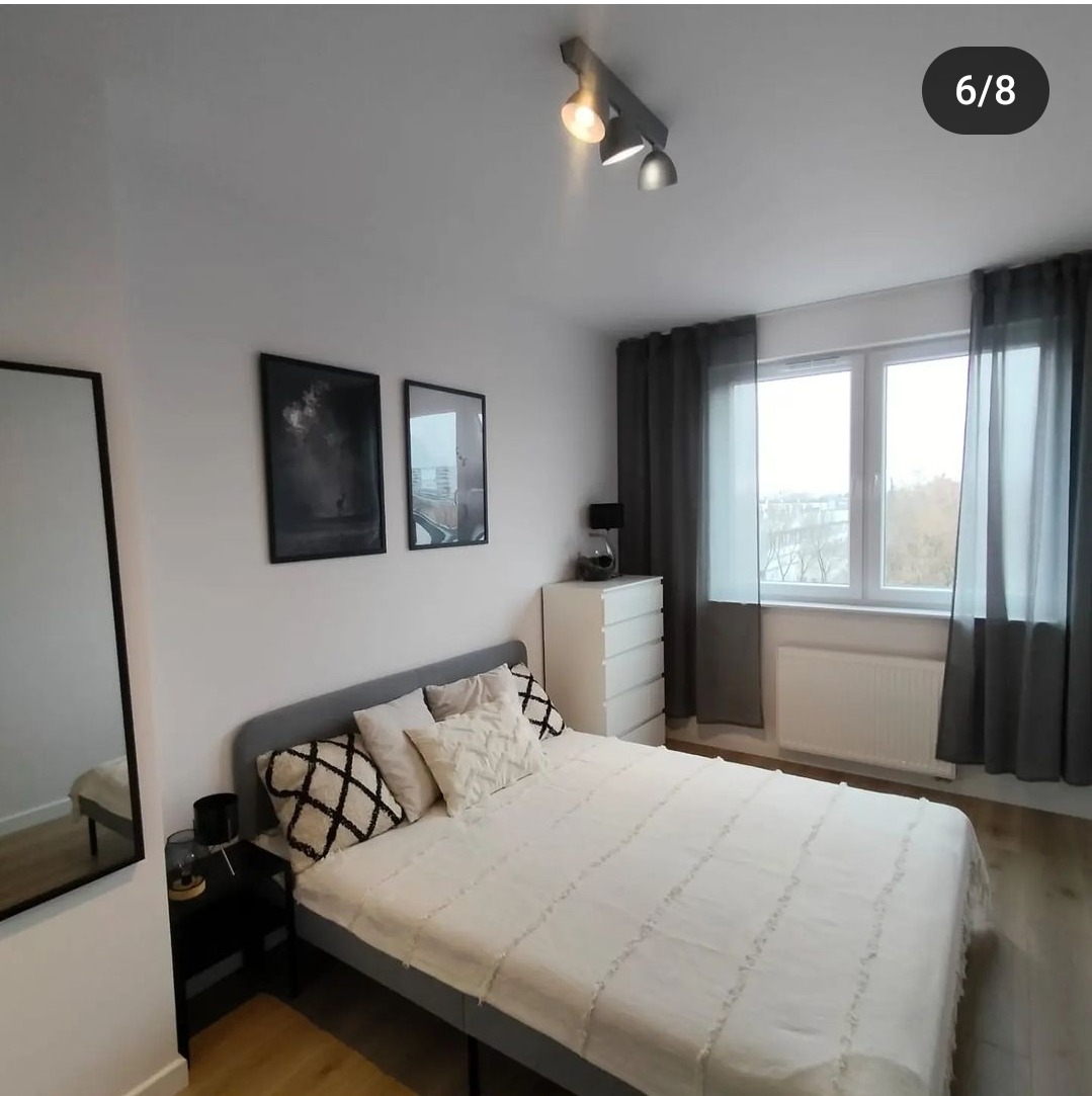 Jasna sypialnia z szarą ramą łóżka, białą pościelą w delikatne paski, wzorzystymi poduszkami, dwoma obrazami na ścianie, białą komodą i szarymi zasłonami przy oknie z widokiem na miasto.