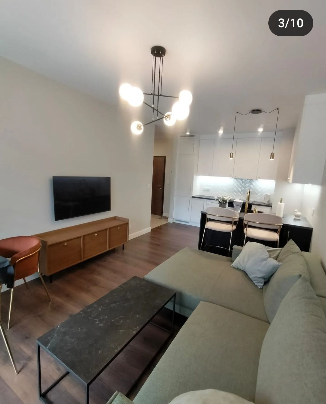 Nowoczesny salon z aneksem kuchennym: szara sofa narożna, czarny stolik kawowy z marmurowym blatem, drewniana szafka RTV pod telewizorem, designerski żyrandol z kulistymi kloszami.