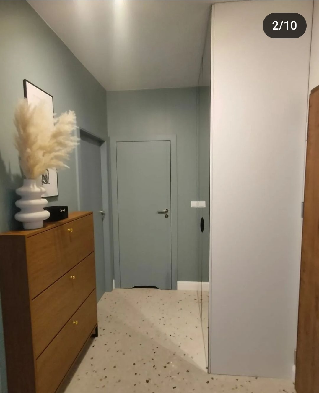 Nowoczesny przedpokój w odcieniach szarości i beżu z drewnianą komodą na buty, ozdobną trawą pampasową w białym wazonie i podłogą lastryko.