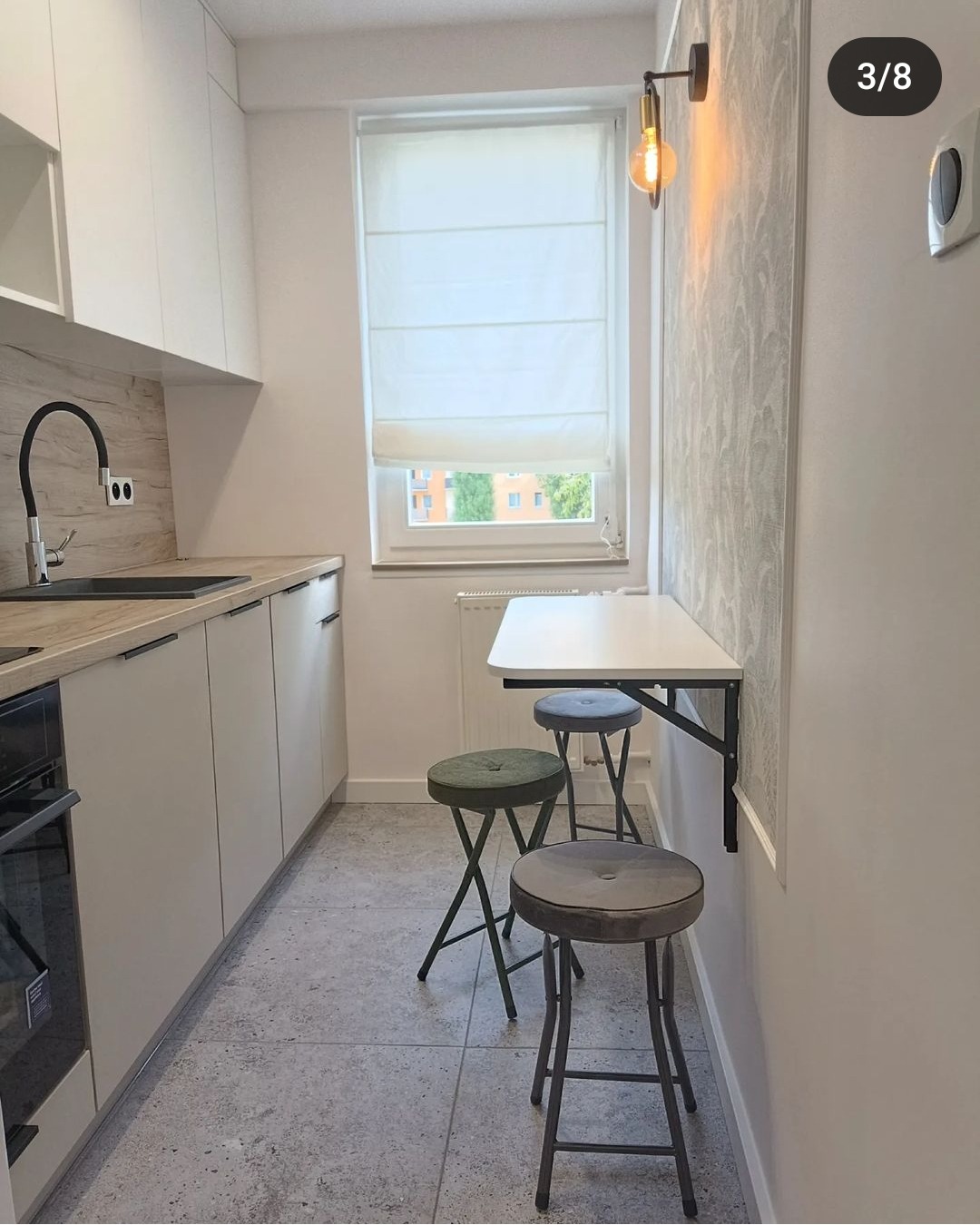 Nowoczesna, jasna kuchnia z białymi szafkami, drewnianym blatem, składanym stolikiem przymocowanym do ściany i dwoma stołkami barowymi z zielonym i szarym siedziskiem, widok na okno z roletą rzymską.