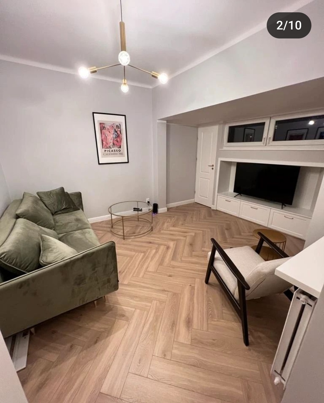 Salon z parkietem ułożonym w jodełkę, zielona sofa, okrągły stolik kawowy, biała zabudowa TV, plakat Picasso na ścianie i nowoczesna lampa sufitowa.