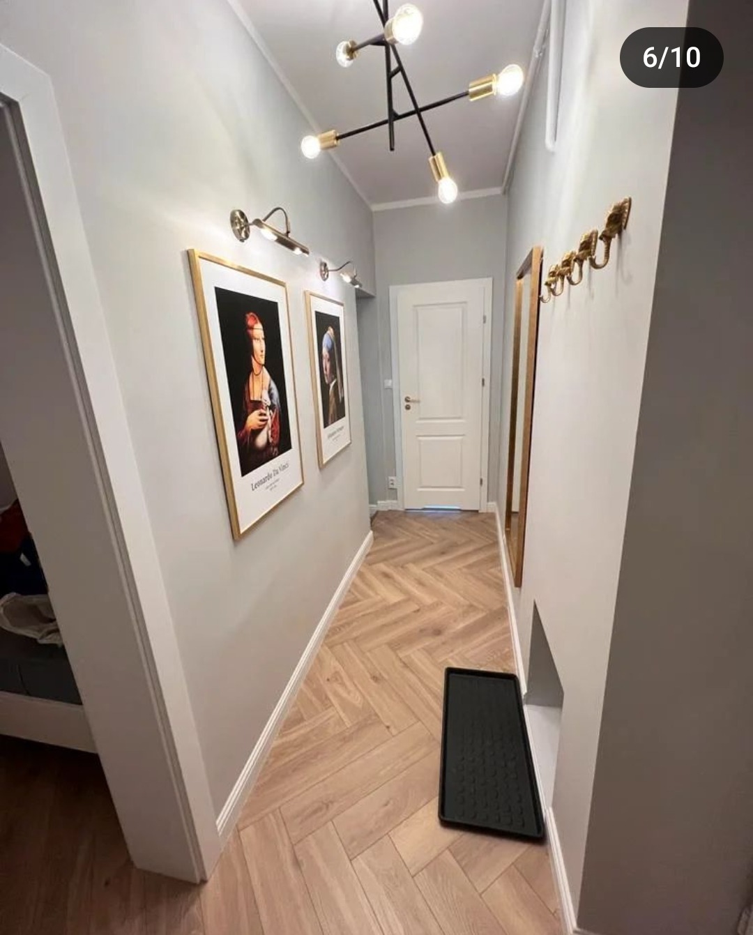 Wąski korytarz z parkietem ułożonym w jodełkę, szare ściany zdobione obrazami w złotych ramach i złotymi kinkietami, nowoczesna lampa sufitowa, lustro w złotej ramie i złote wieszaki.
