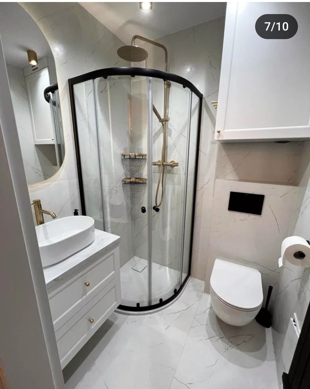 Nowoczesna łazienka z marmurowymi płytkami, kabiną prysznicową z czarną ramą i złotą armaturą, białą umywalką nablatową na szafce z frezowanymi frontami oraz podwieszaną toaletą.