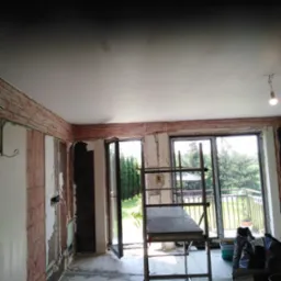 Salon w trakcie remontu: ściany przygotowane pod gładzie, widoczne liczne przewody elektryczne ułożone wzdłuż ścian pod sufitem, rozstawiony metalowy ruszt, otwarte drzwi balkonowe z widokiem...