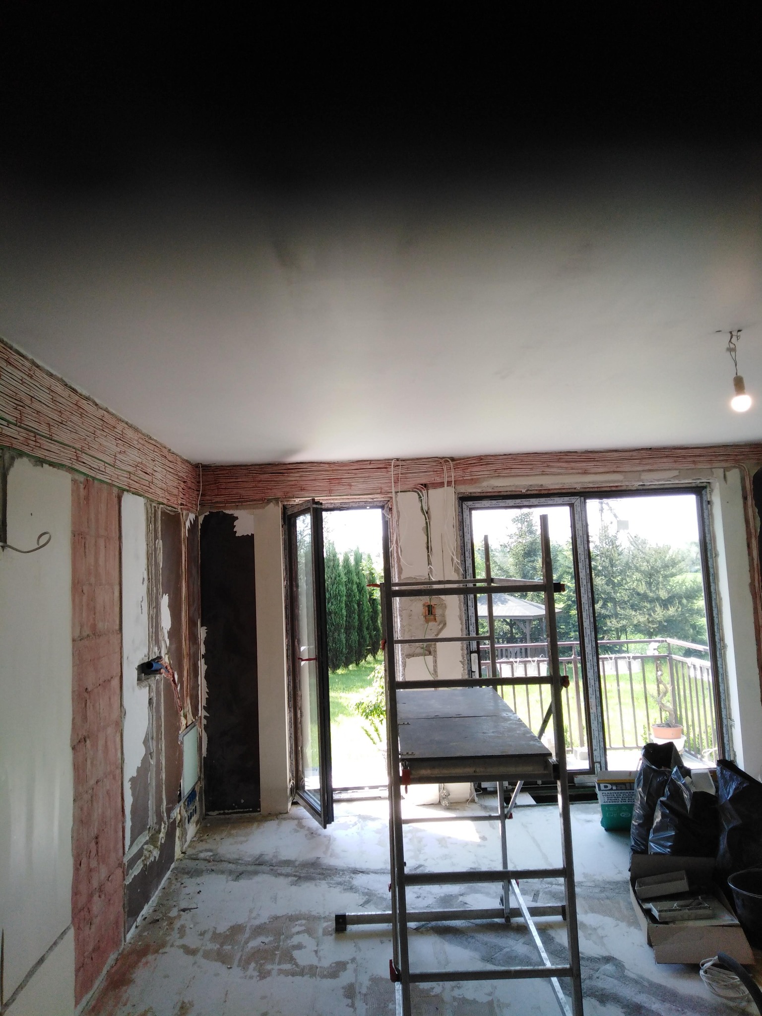 Salon w trakcie remontu: ściany przygotowane pod gładzie, widoczne liczne przewody elektryczne ułożone wzdłuż ścian pod sufitem, rozstawiony metalowy ruszt, otwarte drzwi balkonowe z widokiem...