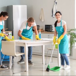 F.U.H ORZEŁ RAFAŁ ZOSGÓRNIK - Trzy osoby w niebieskich fartuchach i żółtych rękawicach sprzątają biały stół w jasnym pomieszczeniu; jedna osoba zamiata podłogę miotłą i szufelką, druga spryskuje powierzchnię, a trzecia wyciera...