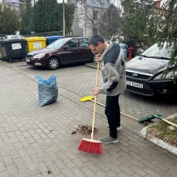 F.U.H ORZEŁ RAFAŁ ZOSGÓRNIK - Mężczyzna zamiata czerwoną szczotką liście na brukowanym podwórku, obok zaparkowane auta i worki na śmieci. Jesienne porządki na zewnątrz budynku.