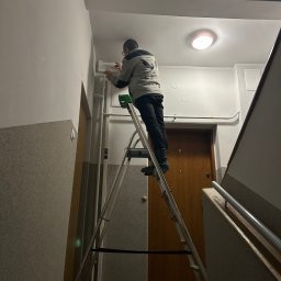 F.U.H ORZEŁ RAFAŁ ZOSGÓRNIK - Pracownik na drabinie montuje listwę przypodłogową w klatce schodowej. Widoczne drzwi wejściowe i fragment schodów. Praca wykonywana wewnątrz budynku.