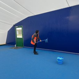 F.U.H ORZEŁ RAFAŁ ZOSGÓRNIK - Pracownik w kamizelce odblaskowej myje niebieską ścianę w hali sportowej za pomocą mopa. Obok stoi wiadro. Widoczna brama wejściowa i biały sufit hali.
