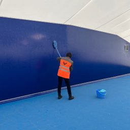 F.U.H ORZEŁ RAFAŁ ZOSGÓRNIK - Pracownik w kamizelce myje niebieską ścianę mopem. Widoczne wiadro z wodą. Hala sportowa lub magazyn. Ślady mycia na ścianie.