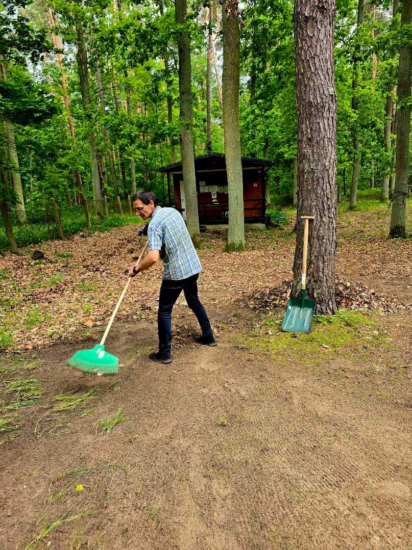 Mężczyzna w lesie zamiata ścieżkę zieloną miotłą. Obok drzewa stoi zielona łopata. W tle widoczny mały budynek.
