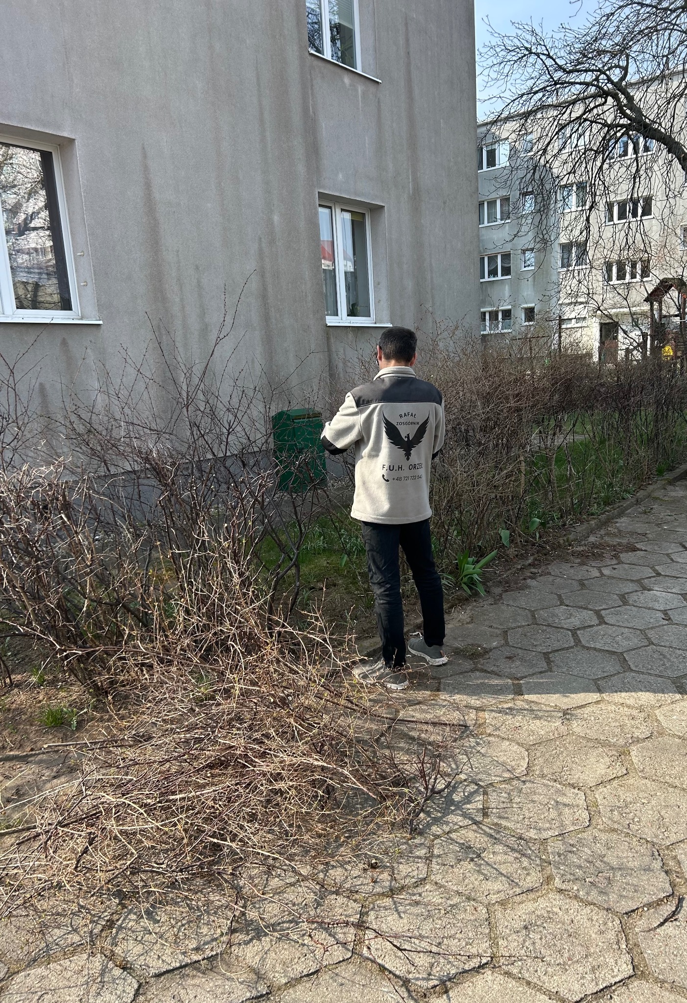 Mężczyzna w firmowej kurtce przycina krzewy przed blokiem. Widoczne ścięte gałęzie na chodniku. Praca w toku, słoneczny dzień.