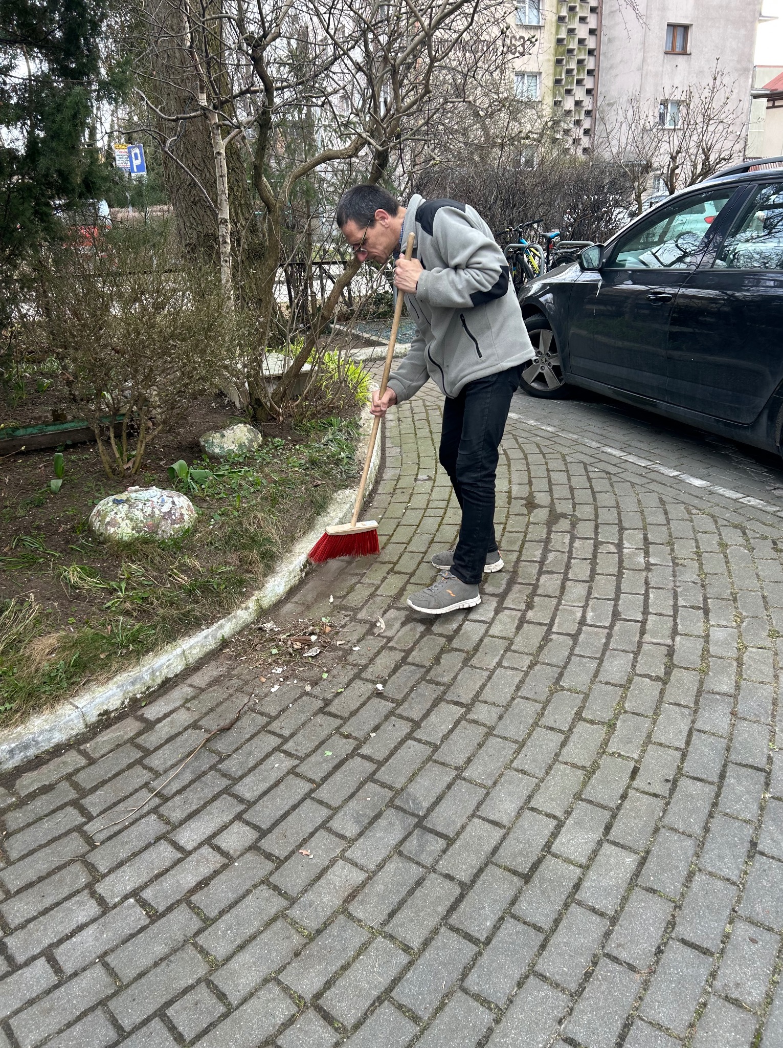 Mężczyzna w okularach zamiata chodnik z kostki brukowej czerwoną szczotką. W tle zaparkowany samochód i budynek mieszkalny.