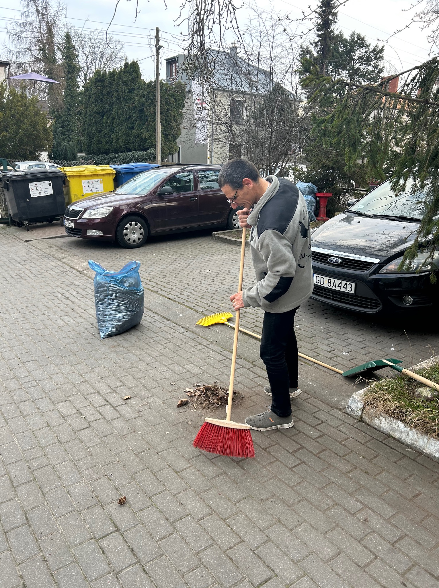Mężczyzna zamiata czerwoną szczotką liście na brukowanym podwórku, obok zaparkowane auta i worki na śmieci. Jesienne porządki na zewnątrz budynku.