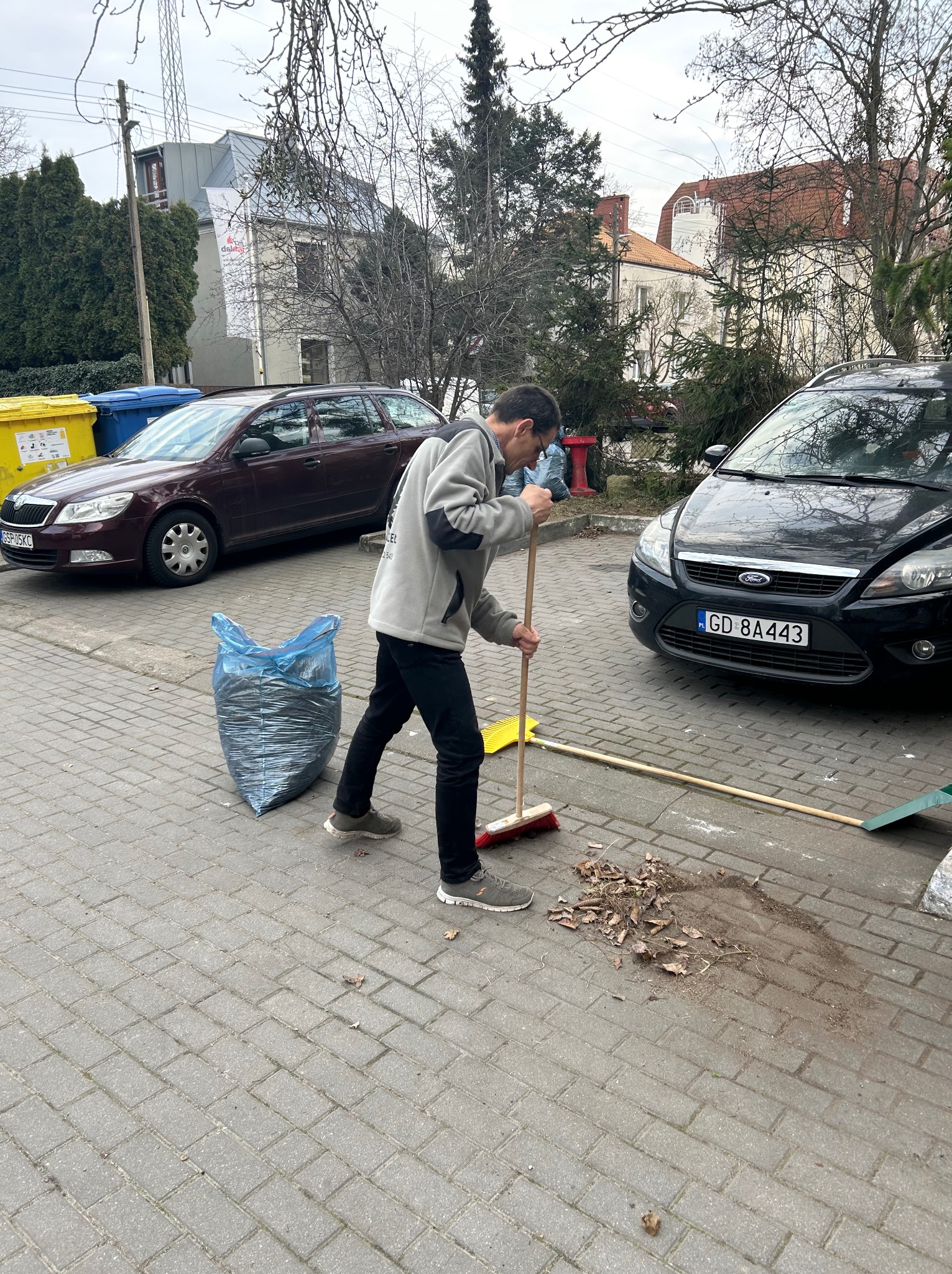 Mężczyzna zamiata liście na chodniku przed domem, obok zaparkowane auta i worki na śmieci. Jesienne porządki na posesji.