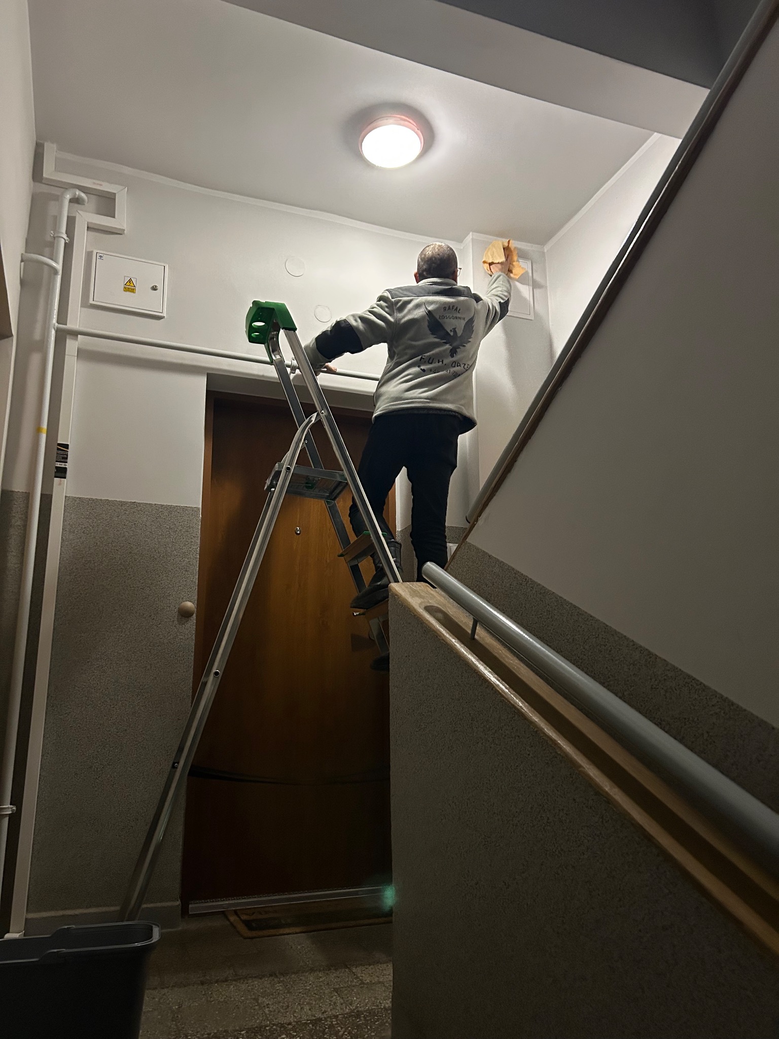 Pracownik na drabinie czyści ścianę na klatce schodowej, używając ściereczki. Widoczne rury, skrzynka elektryczna i fragment schodów. Ujęcie z dołu.