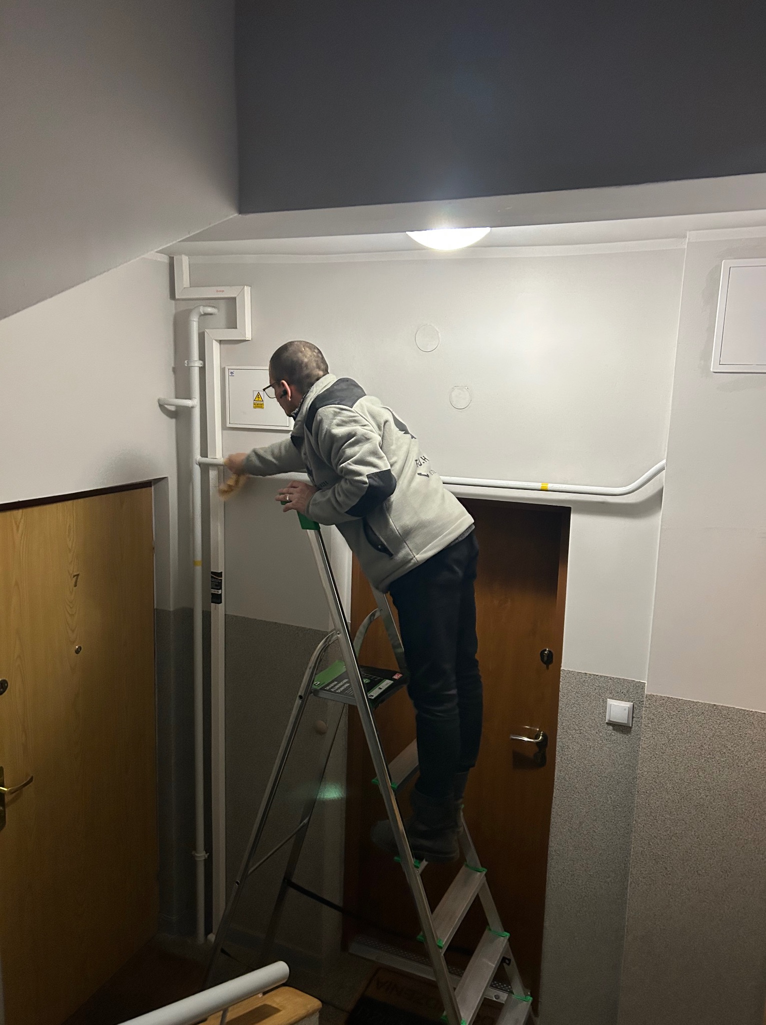 Mężczyzna na drabinie czyści rury w korytarzu budynku. Widoczne drzwi z numerem 7, białe ściany i szara góra. Praca wykonywana z użyciem ściereczki.