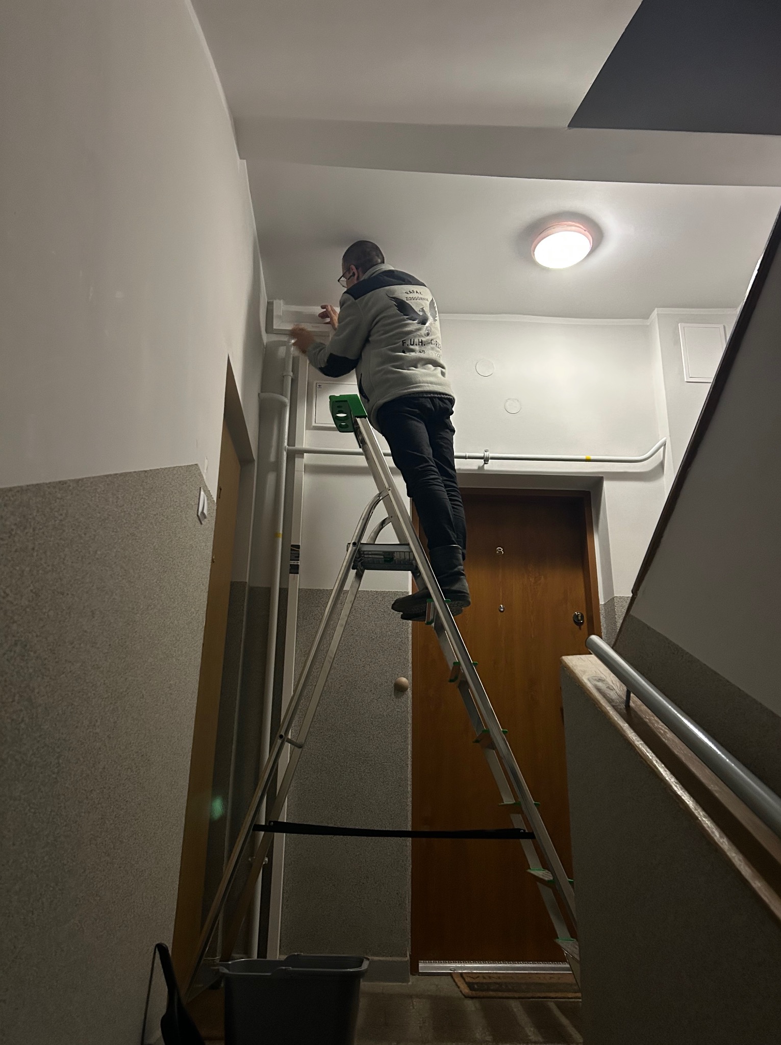 Pracownik na drabinie montuje listwę przypodłogową w klatce schodowej. Widoczne drzwi wejściowe i fragment schodów. Praca wykonywana wewnątrz budynku.